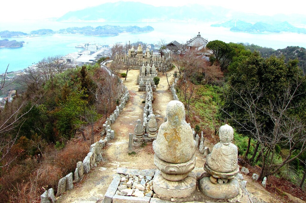山道の両脇に並ぶ白い石仏の列と、奥に広がる海